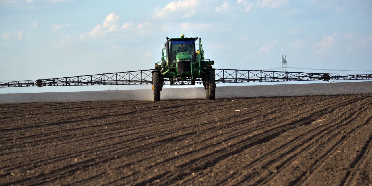 A green tractor equipped with a large spraying mechanism is applying substances to a dry, plowed field. The tractor is positioned in the foreground, facing the viewer, with a clear sky and distant structures visible in the background.