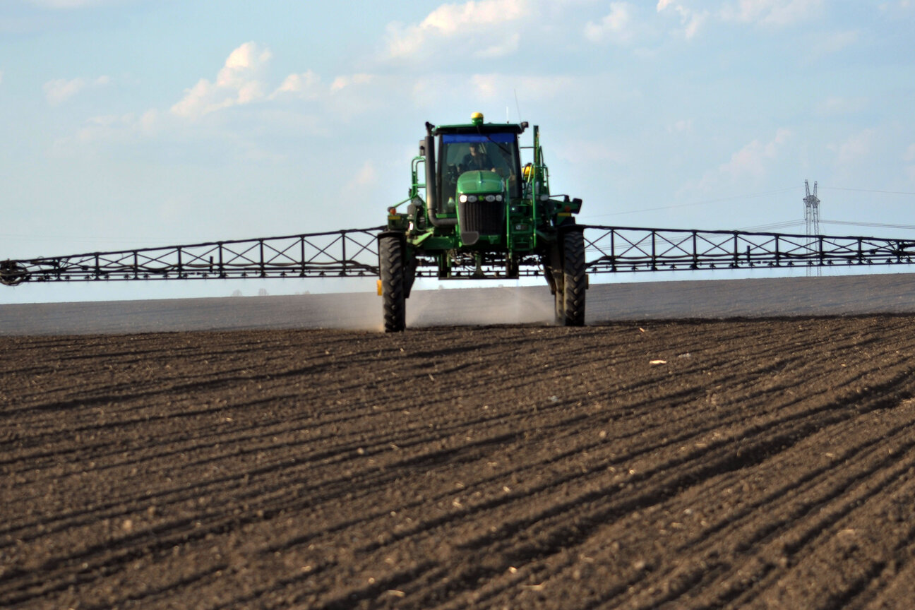 A green tractor equipped with a large spraying mechanism is applying substances to a dry, plowed field. The tractor is positioned in the foreground, facing the viewer, with a clear sky and distant structures visible in the background.