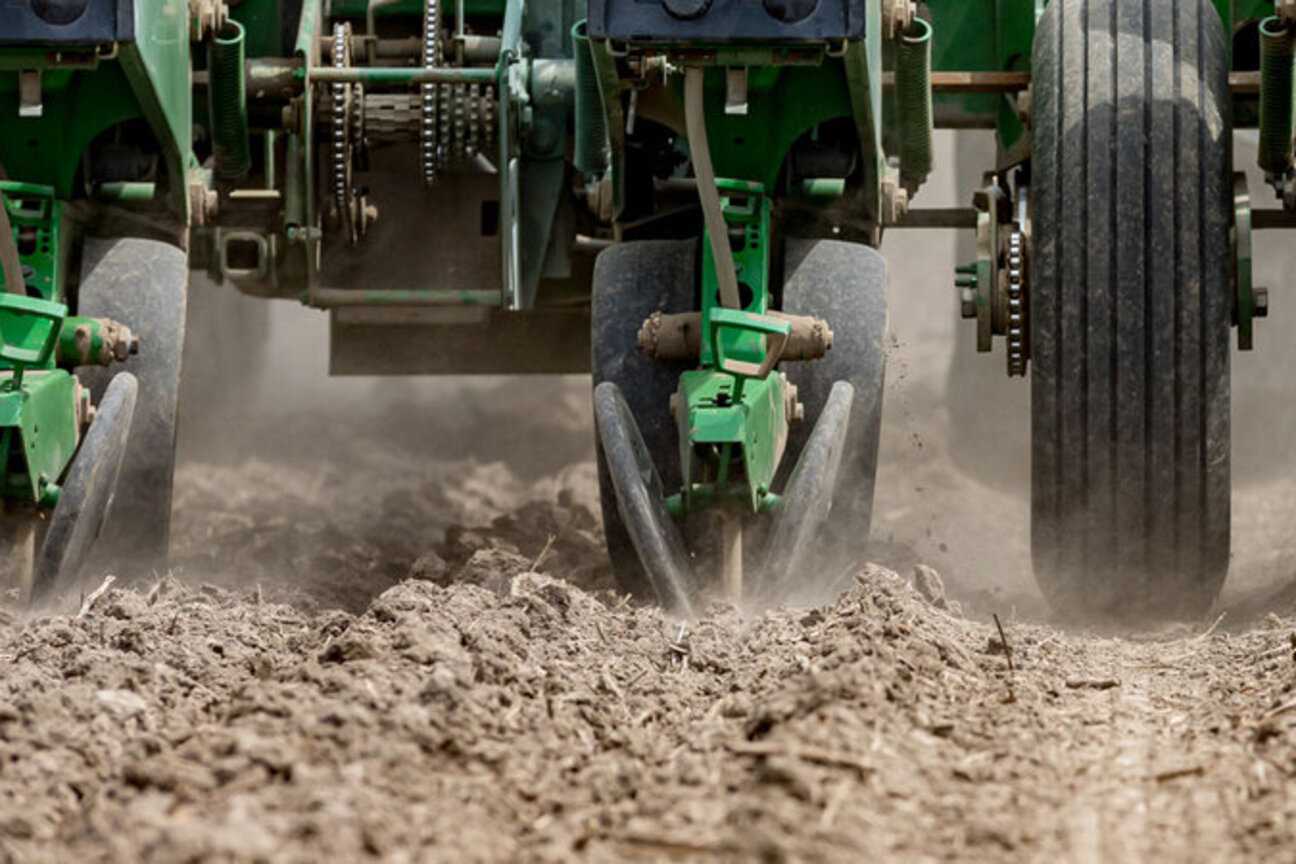 The image shows the wheels of a farming machinery planting in freshly tilled soil. Dust is being kicked up as the equipment moves forward, indicating active agricultural work. The green wheels contrast against the brown earth.