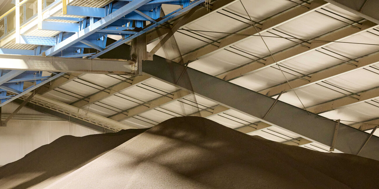 A large pile of dark material is seen inside a spacious industrial building with a high ceiling. Above the pile, there is a blue overhead structure, suggesting an area for loading or processing materials. The setting appears to be a warehouse or manufacturing facility.