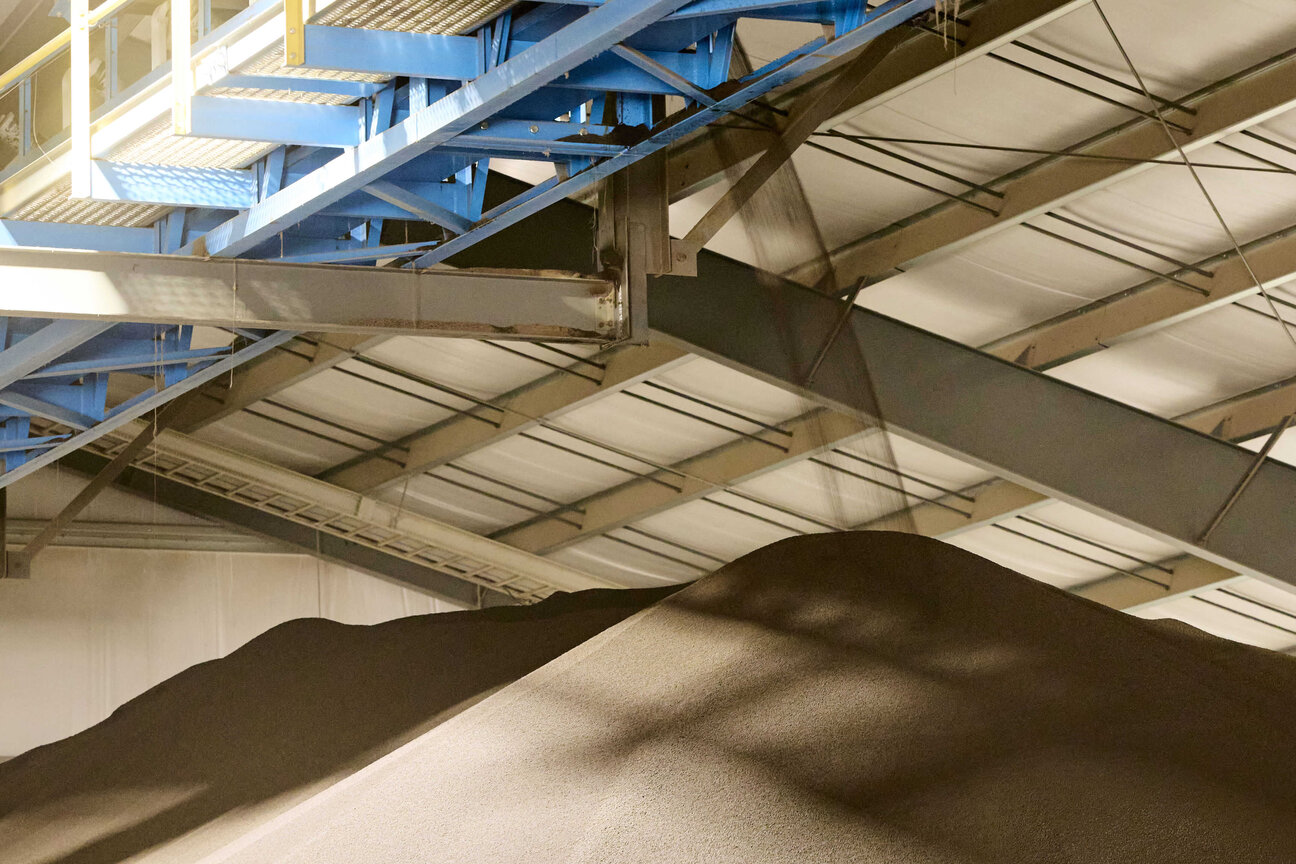 A large pile of dark material is seen inside a spacious industrial building with a high ceiling. Above the pile, there is a blue overhead structure, suggesting an area for loading or processing materials. The setting appears to be a warehouse or manufacturing facility.