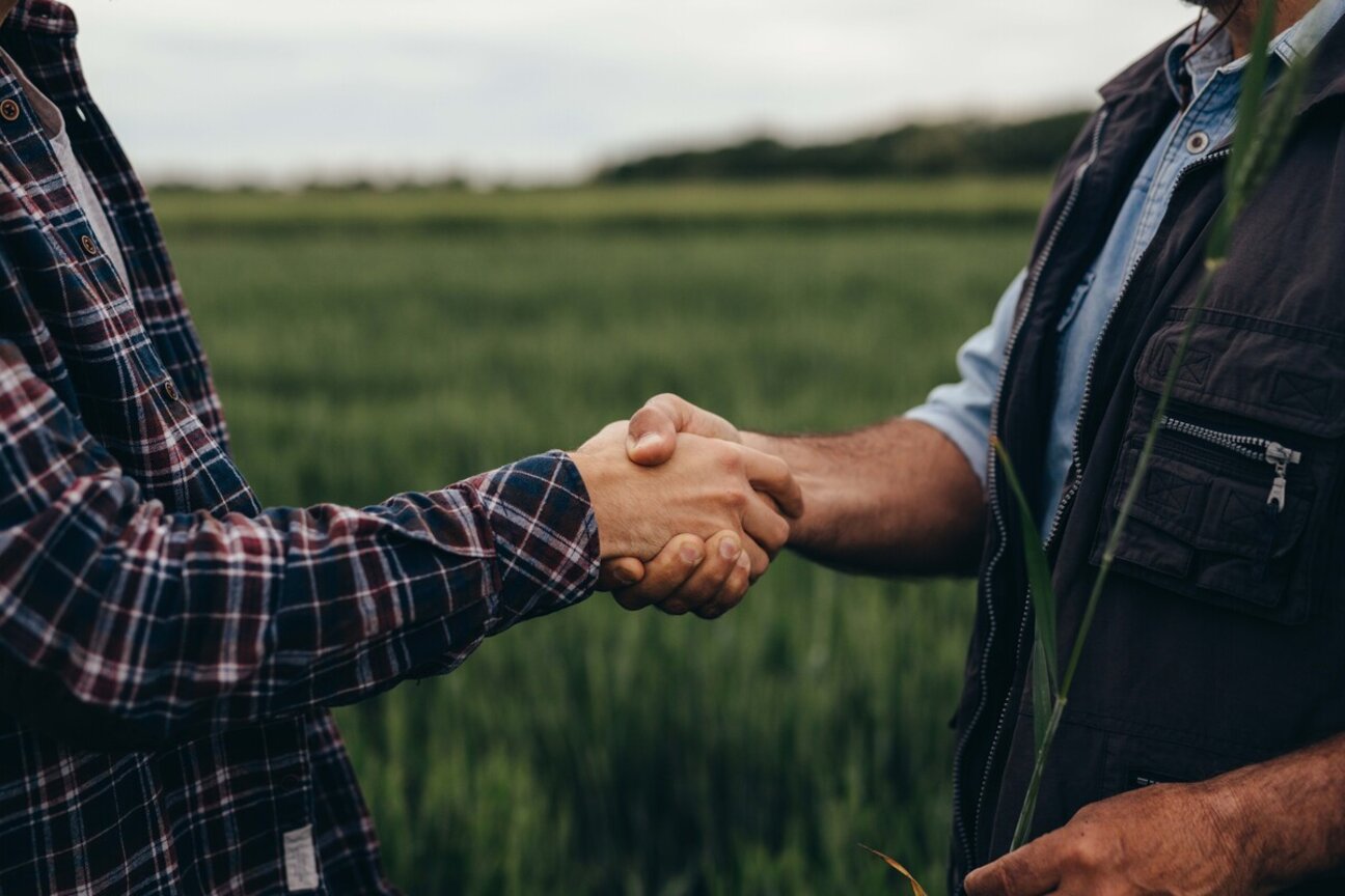 Two people are shaking hands in a green field. One is wearing a plaid shirt, and the other is dressed in a sleeveless vest over a shirt. The handshake signifies a partnership or agreement, set against a backdrop of lush vegetation.