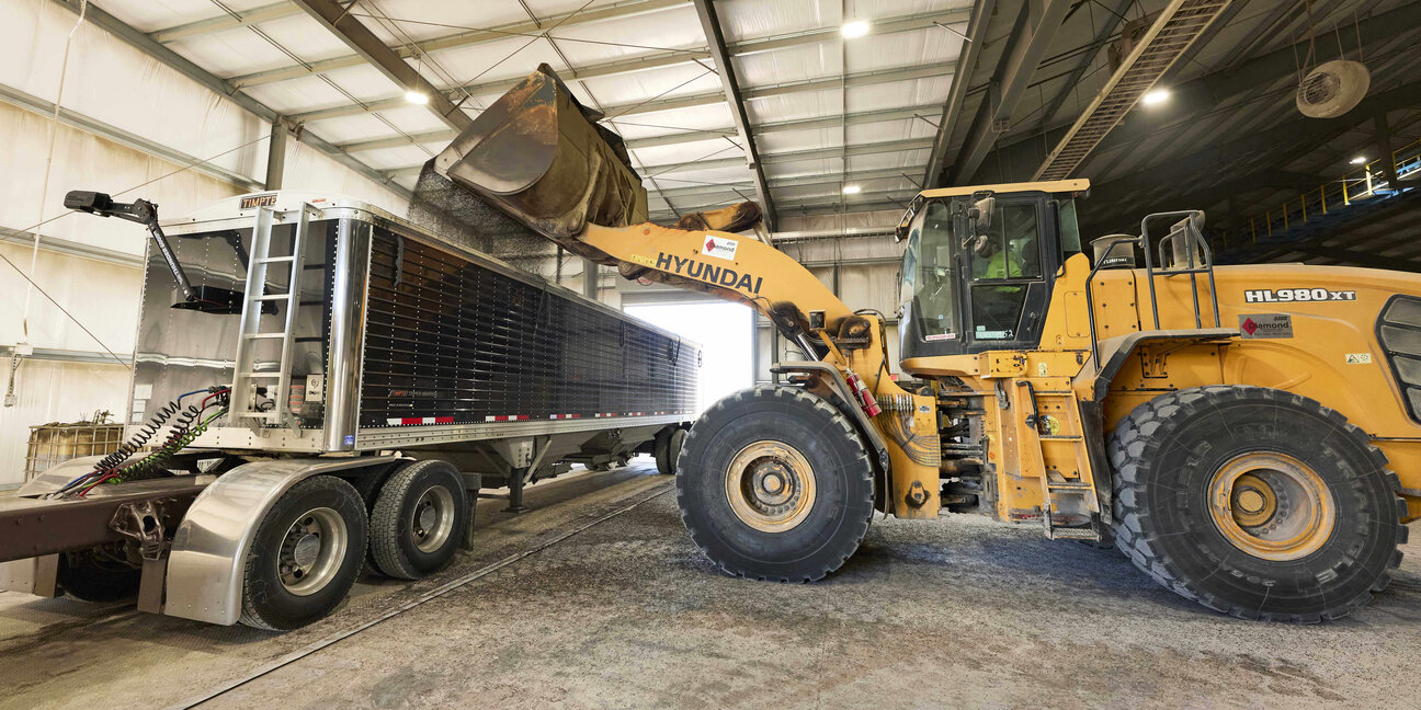 A large yellow loader is positioned beside a semi-truck, using its bucket to load material into the truck's trailer. The scene is set indoors, within a warehouse-like environment, with high ceilings and visible structural elements.