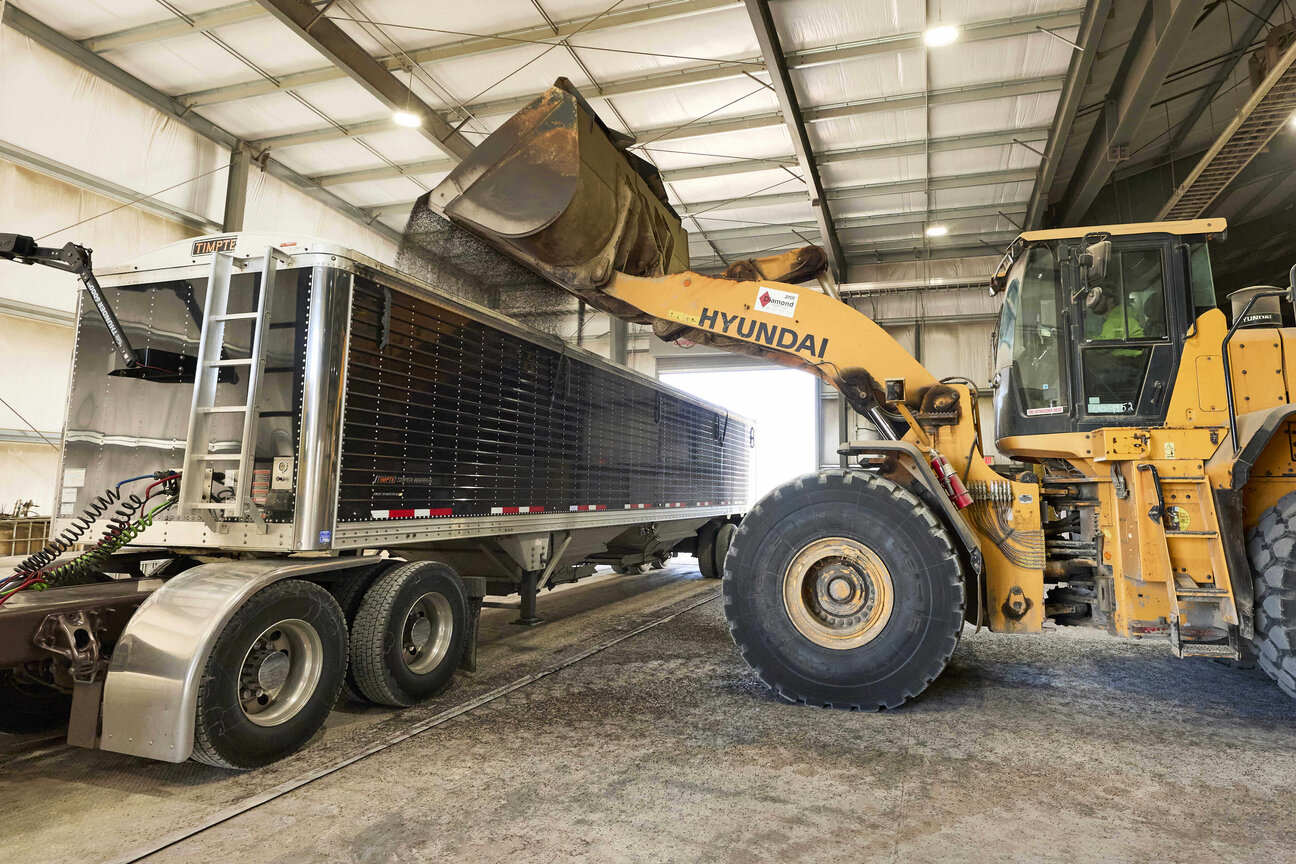 A large yellow loader is positioned beside a semi-truck, using its bucket to load material into the truck's trailer. The scene is set indoors, within a warehouse-like environment, with high ceilings and visible structural elements.