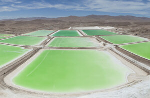 A panoramic view of salt flats divided into large rectangular ponds filled with bright green water. The landscape is arid and mountainous, with a clear blue sky above. The vibrant green contrasts with the white salt and barren earth surrounding the ponds.
